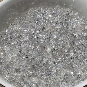 May include: A white teacup with a gold rim filled with clear, sparkling crystals. The teacup is sitting on a white saucer.