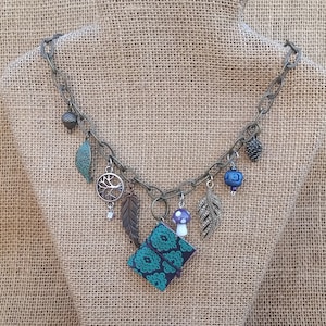 Puede incluir: Un collar de cadena plateada con múltiples dijes, incluyendo un árbol, hojas, un hongo y un pequeño libro con una cubierta de patrón verde azulado y verde.