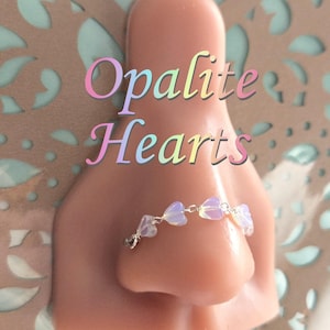 May include: A nose ring featuring small, opalescent heart-shaped beads strung on a delicate silver chain. The text "Opalite Hearts" is displayed in a soft, multi-colored font above the nose ring.
