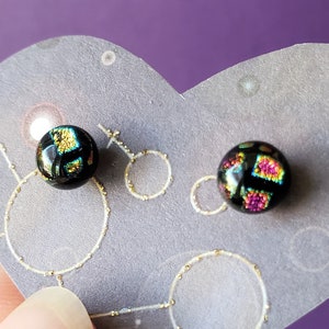 May include: A pair of black stud earrings with iridescent glass insets. One earring has a gold and green pattern, the other has a pink and purple pattern.