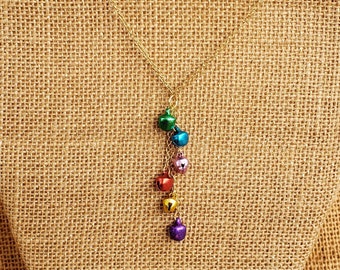 Puede incluir: Un collar de cadena dorada con un dije colgante de campanillas de colores. Las campanillas son verdes, azules, rosas, naranjas y moradas.
