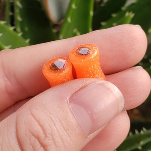 May include: A pair of orange and brown glitter ear gauges. The gauges are cylindrical and have a flat, circular top with a brown glitter design.