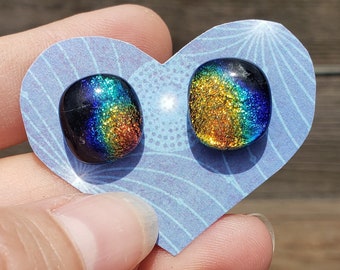 May include: A pair of iridescent glass earrings with a rainbow effect. The earrings are oval shaped and have a dark blue base with a gold and green iridescent sheen.