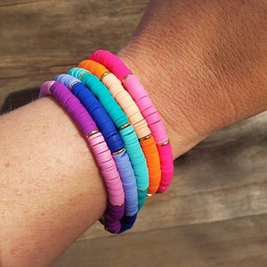 May include: A stack of colorful bracelets made of round beads with gold accents. The bracelets are layered on a person's wrist.