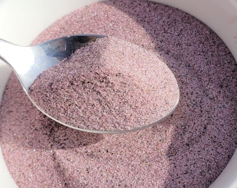 May include: A white teacup filled with a pinkish-purple granular substance, with a silver spoon scooping up a portion. The cup has a gold rim and sits on a white saucer with a honeycomb pattern. The background is a wooden surface.
