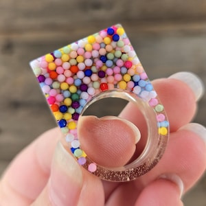 May include: A clear resin ring with a square top filled with colorful, tiny spheres. The ring band is clear, and the square top features a variety of colors, including pink, yellow, blue, and green. The ring is a unique, eye-catching accessory.