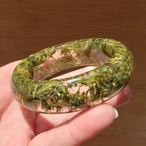 Puede incluir: Un brazalete de resina transparente con material vegetal verde encapsulado. El brazalete de forma ovalada tiene un acabado liso y pulido. El material vegetal verde es visible en todo el brazalete, creando un aspecto natural único. El brazalete se sostiene en una mano.