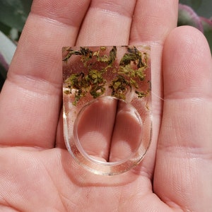 May include: A clear resin ring with dried green foliage embedded inside. The ring is shown being held by a hand.
