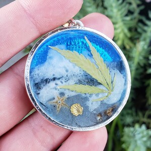 Puede incluir: Un colgante de plata con un diseño de resina azul, blanco y gris. La resina contiene una hoja verde prensada, una concha y una estrella de mar.