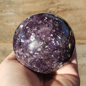 Peut inclure: Une sphère de quartz de Lemurie de couleur violette et argentée. La sphère est lisse et polie, avec un motif moucheté d'argent et de violet.
