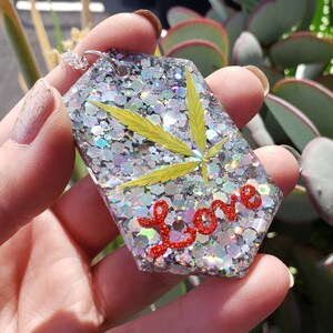May include: A silver glitter resin pendant with a pressed green leaf and the word "Love" in red glitter. The pendant is on a silver chain.
