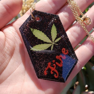 May include: A black resin pendant necklace with a gold chain. The pendant features a pressed green leaf and the word "Fire" in red glitter.