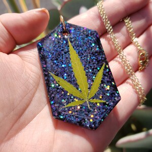 May include: A gold chain necklace with a purple and iridescent glitter resin pendant featuring a pressed green leaf.