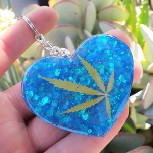 May include: Heart-shaped keychain with a blue glitter background and a green cannabis leaf. The keychain is attached to a silver metal ring. The heart is made of resin.