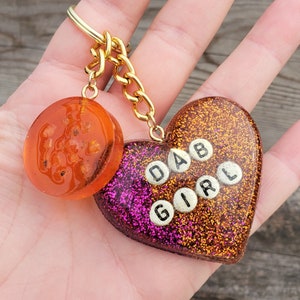 Puede incluir: Un llavero de oro con un dije en forma de corazón que dice "DAB GIRL" en letras dorées sobre un fondo de purpurina rosa y naranja. El llavero también tiene un dije redondo naranja con una textura brillante.
