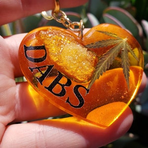 Puede incluir: Un llavero de resina en forma de corazón con una cadena dorada, una hoja de marihuana verde y la palabra "DABS" en letras negras. La resina es transparente y de color ámbar, y hay un pequeño trozo de resina de color dorado en el interior.