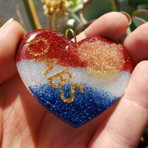 Puede incluir: Adorno de resina en forma de corazón rojo, blanco y azul con purpurina dorada y la palabra "BABS" escrita en purpurina dorada.