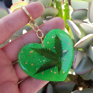 Puede incluir: Un llavero en forma de corazón verde con una cadena dorada y una hoja prensada en su interior. La hoja es una hoja de marihuana y el corazón está cubierto de purpurina.