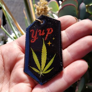 以下が含まれることがあります： 緑のマリファナの葉と赤いグリッターで「Yup」と書かれた黒いレジン製のキーホルダーです。