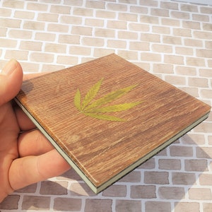 May include: A square wooden coaster with a green leaf design. The coaster has a light brown wood grain and a dark brown border.