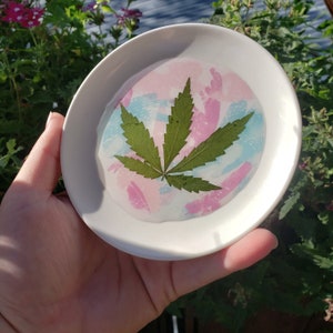 May include: A white ceramic dish with a pink, blue, and white swirl design. A green cannabis leaf is pressed into the resin, creating a unique and decorative piece.