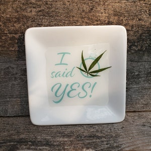 May include: A square white ceramic trinket dish featuring the light blue text I said YES! and a graphic of a diamond ring. A small green cannabis leaf is placed over the ring graphic on the center of the smooth, glossy white surface.