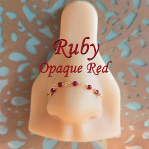 Puede incluir: Un aro de nariz dorado con pequeñas cuentas redondas de color rubí. El aro de nariz se muestra en una nariz de maniquí. El texto "Ruby Opaque Red" está escrito en rojo sobre el aro de nariz.