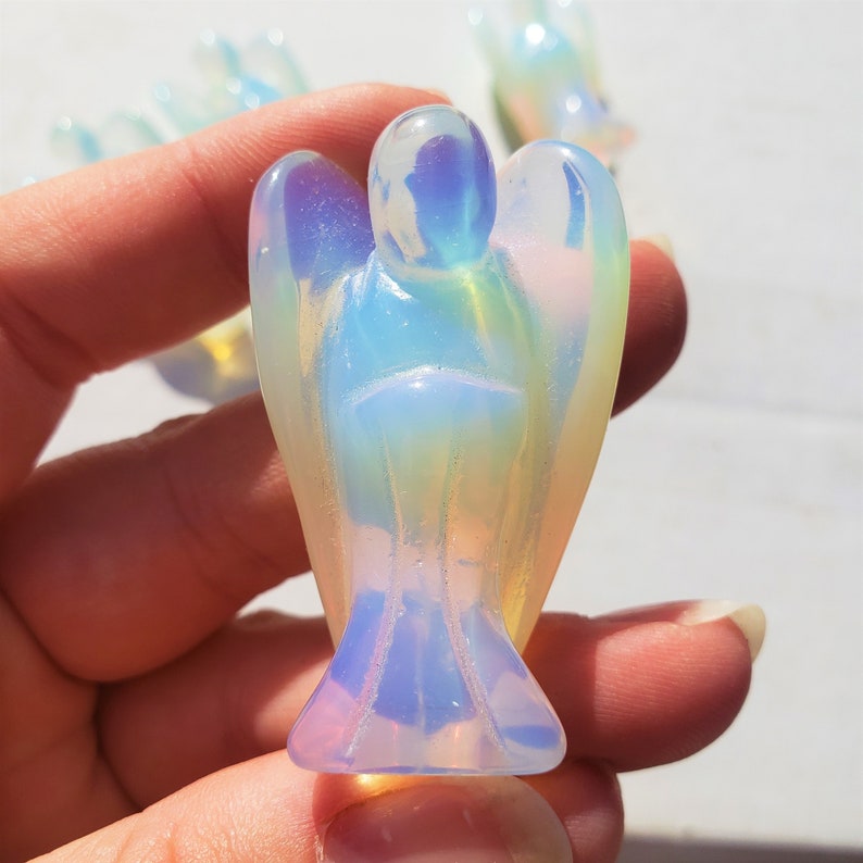 May include: A small, iridescent opal angel figurine with a white, blue, and yellow color scheme. The angel is carved with wings, a head, and a body. The figurine is held in a hand.