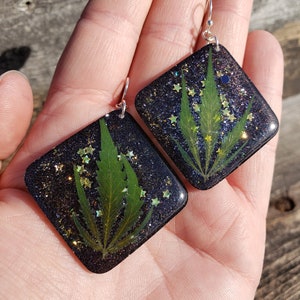 Puede incluir: Un par de pendientes de resina negra con ganchos plateados. Cada pendiente presenta una hoja verde prensada con purpurina dorada incrustada en la resina.