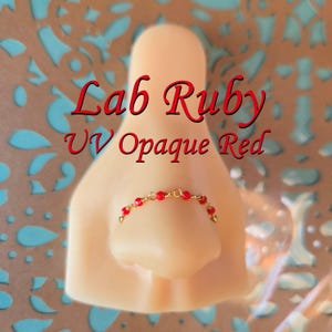 May include: A nose ring with red beads and gold-colored wire is displayed on a mannequin nose. The text "Lab Ruby UV Opaque Red" is written in red script above the nose ring.