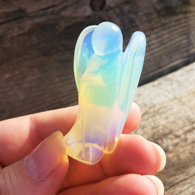 May include: A small, iridescent angel figurine carved from opalite. The angel is in a praying position with its wings folded behind it. The figurine is a pale yellow, blue, and pink color.