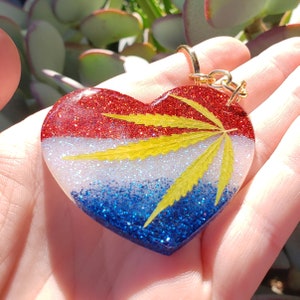 May include: Heart-shaped keychain with red, white, and blue glitter, and a yellow cannabis leaf. The resin pendant has a gold-colored metal keyring. The design is reminiscent of a flag.