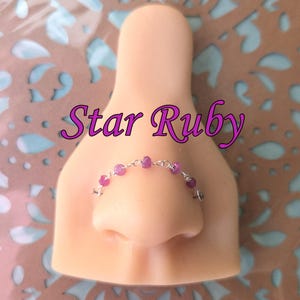 May include: A nose ring featuring small, round, pink gemstones on a silver chain. The text "Star Ruby" is written above the nose ring in a cursive font. The nose ring is displayed on a mannequin nose.