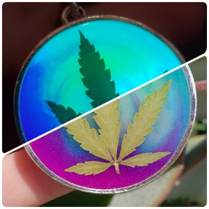 May include: A silver pendant with two resin-filled circles. One circle features a green marijuana leaf on a blue and green background. The other circle features a dried marijuana leaf on a purple and blue background.