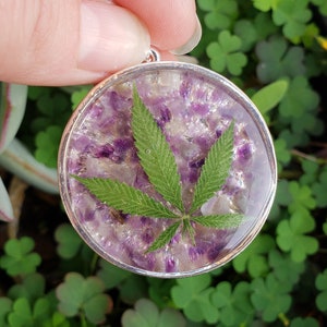 Puede incluir: Un colgante de plata con una cúpula de resina transparente que contiene una mezcla de piedras trituradas de color púrpura y blanco y una hoja verde.