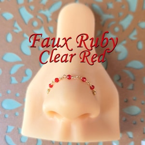 May include: A gold-coloured nose chain with clear red beads, displayed on a mannequin nose. The text "Faux Ruby Clear Red" is written in red script above the nose chain.