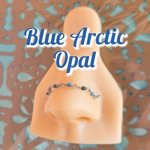 May include: A nose ring made of silver-colored metal with small blue and teal beads. The text "Blue Arctic Opal" is written in a blue script font above the nose ring.