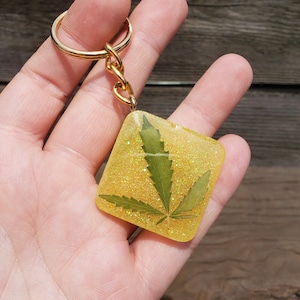 Puede incluir: Un llavero cuadrado dorado con una hoja de cannabis encapsulada en resina transparente. La resina tiene un aspecto amarillo brillante. El llavero tiene un anillo y una cadena dorados.