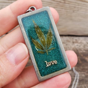 Puede incluir: Un collar con colgante rectangular con un marco plateado. El colgante presenta un fondo verde brillante con una hoja de cannabis seca y la palabra "love" en letras plateadas. El collar lo sostiene una persona.
