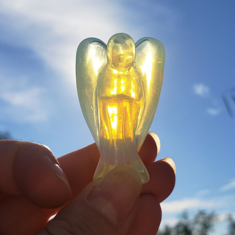 May include: A yellow and green glass angel figurine with wings. The angel is holding a small, clear glass object in its hands.