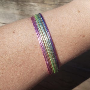 Puede incluir: Un conjunto de pulseras finas en colores vibrantes: morado, verde y azul. Las pulseras brillantes están apiladas en un brazo. Las pulseras parecen estar hechas de un material reflectante.