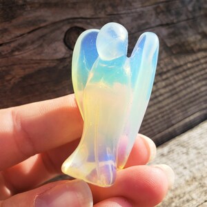 May include: A small, iridescent angel figurine carved from opalite. The angel has a rounded head and outstretched wings. The figurine is a pale yellow color with a rainbow sheen.