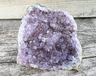 May include: A large amethyst crystal cluster with a natural, rough exterior and a purple, sparkly interior. The crystal is sitting on a wooden surface.