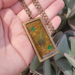 Puede incluir: Un collar dorado con un colgante rectangular que presenta un trébol de cuatro hojas verde y una hoja verde prensada en resina de purpurina dorada.