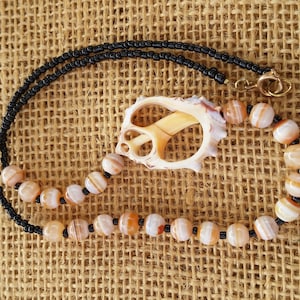 Puede incluir: Un collar con un colgante de concha y cuentas de ágata marrón y blanca alternas. El collar está enhebrado en hilo negro.