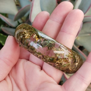 May include: A clear resin bangle bracelet with embedded dried flowers and gold leaf. The bracelet is a light pink color and has a natural, organic look.