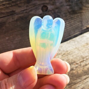 May include: A small, iridescent opal angel figurine with a white, blue, and yellow color scheme. The angel is carved in a standing pose with wings outstretched.