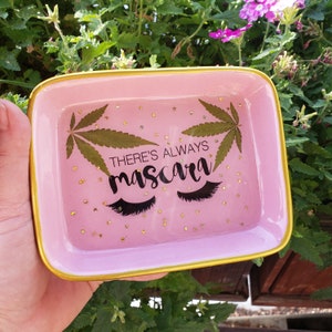 May include: Pink rectangular tray with a gold rim. The tray has a glittery pink surface with gold glitter stars and two black eyelashes. The tray also has a green leaf and the text "THERE'S ALWAYS mascara".