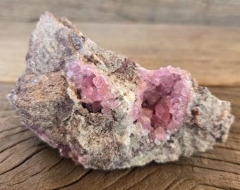 May include: A raw pink crystal cluster with two cavities filled with pink crystals. The crystal cluster is on a wooden surface.