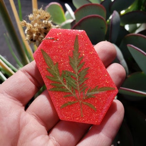 Puede incluir: Un llavero hexagonal de resina roja con purpurina con un helecho verde prensado en su interior.